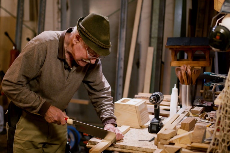 Un homme scie du bois dans un atelier avec une scie à main, entouré d'outils et de matériaux.