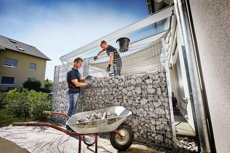 Deux hommes remplissent une clôture de gabions avec des pierres provenant d'une brouette.