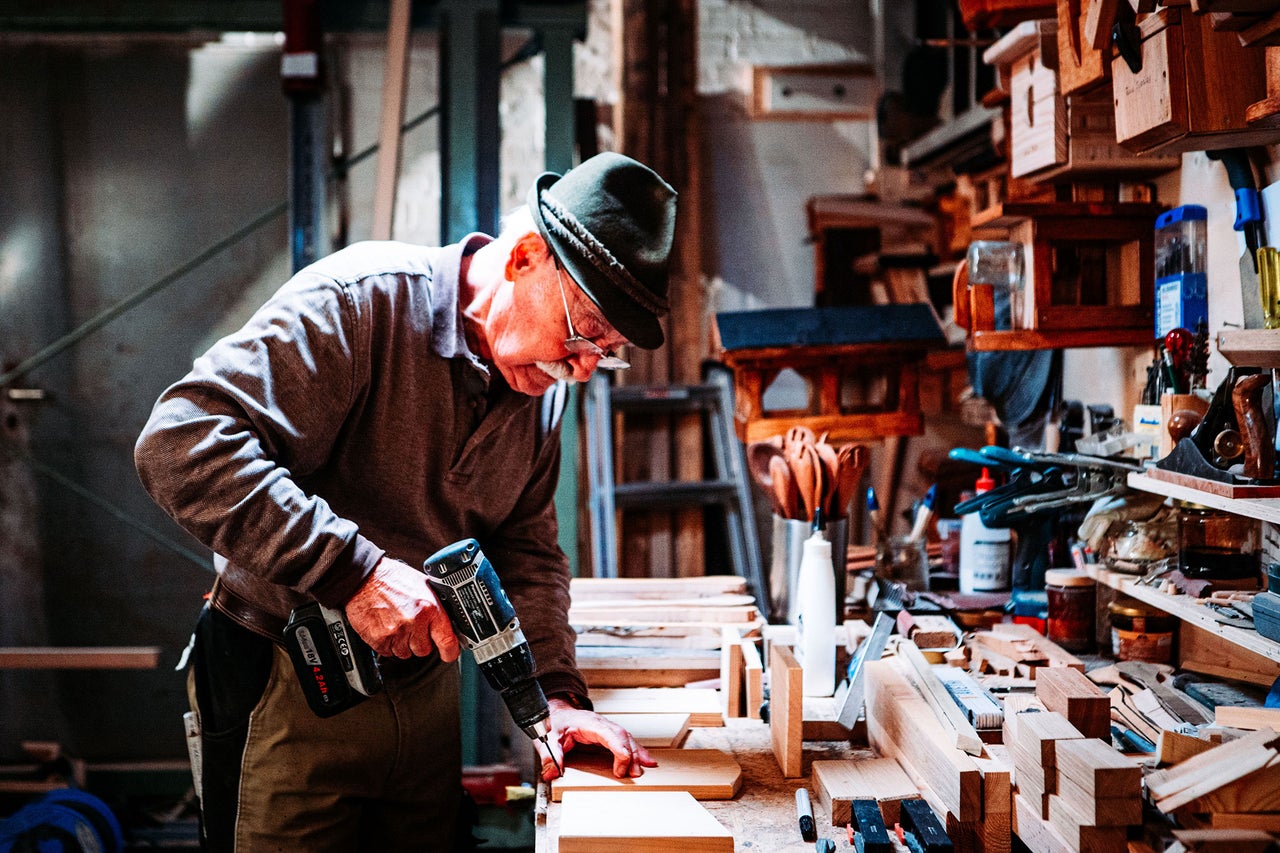 Un homme coiffé d'un chapeau perce du bois avec une visseuse sans fil dans un atelier rempli de nichoirs et d'outils.