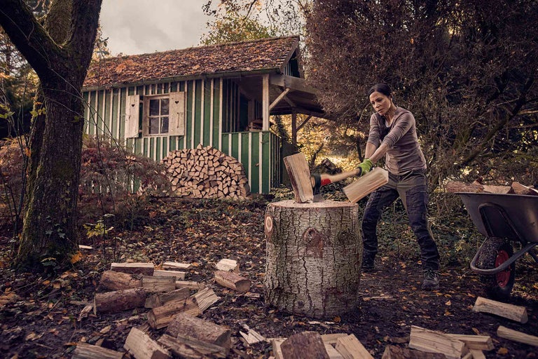 Une femme fend du bois avec une hache devant une remise à bois dans un jardin.