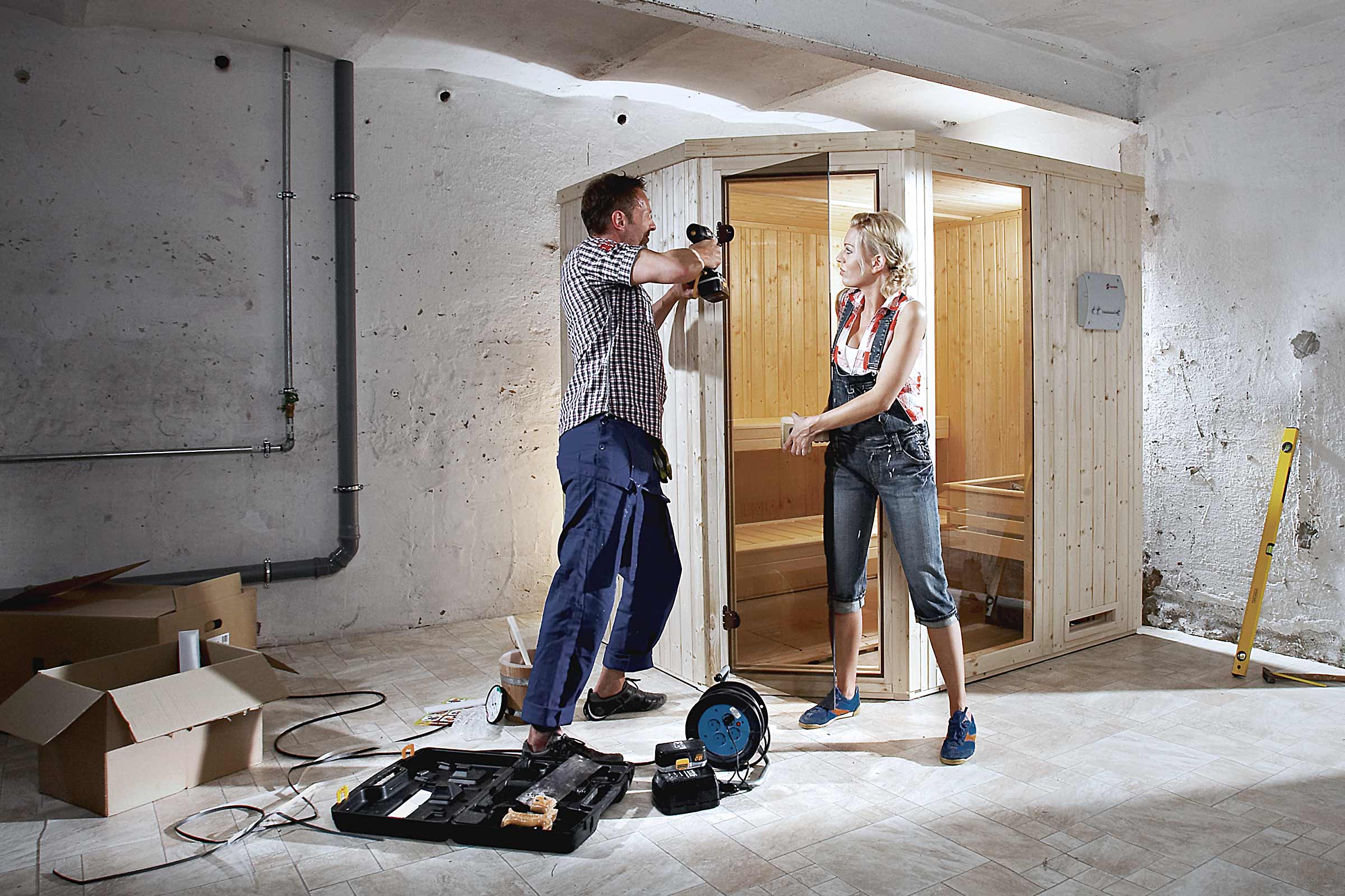 Mann und Frau bauen eine Holzsauna im Keller auf. Werkzeuge und Baumaterialien sind im Raum verteilt.