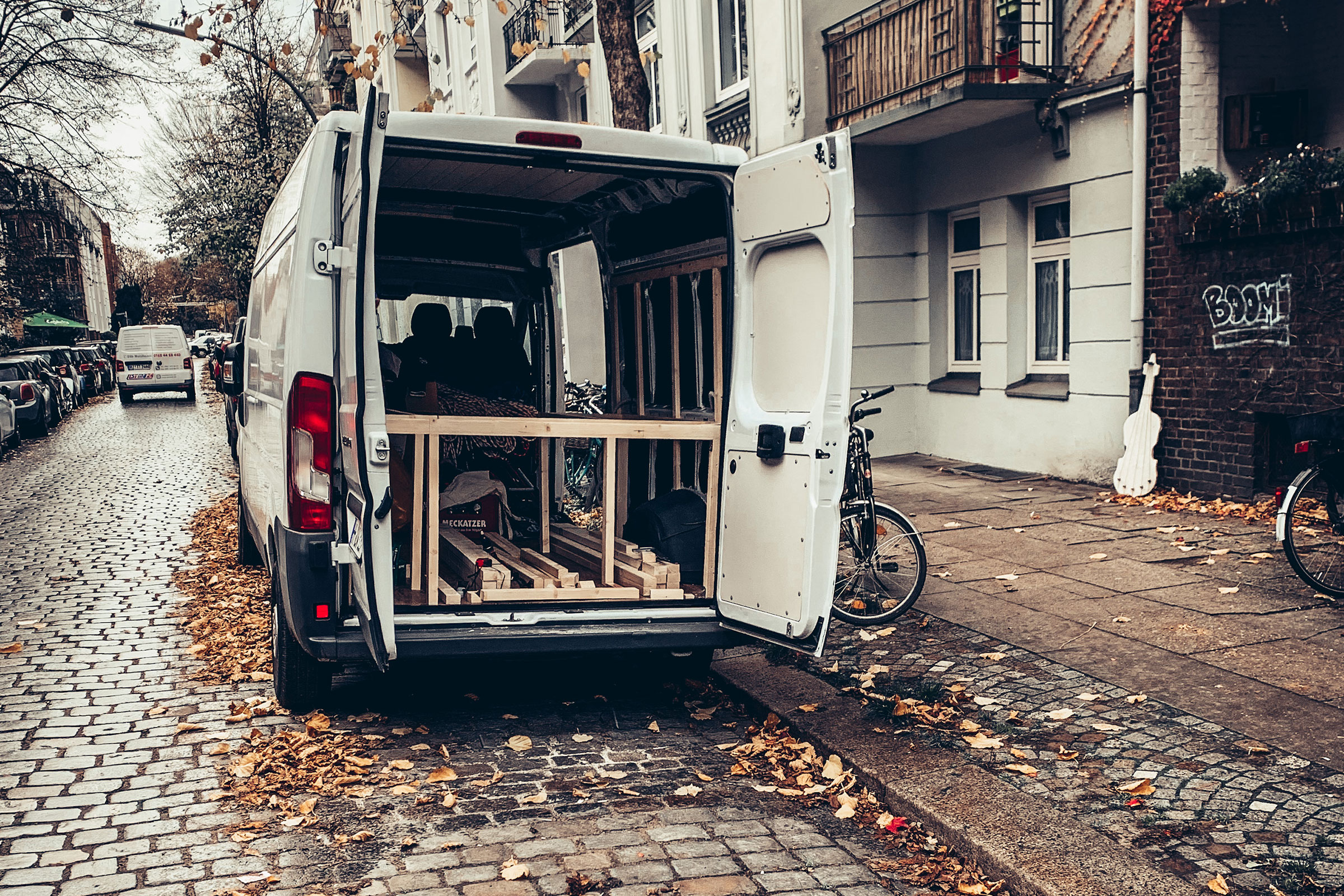 Ein weißer Transporter mit geöffneten Hecktüren auf einer Straße mit Kopfsteinpflaster und Herbstlaub