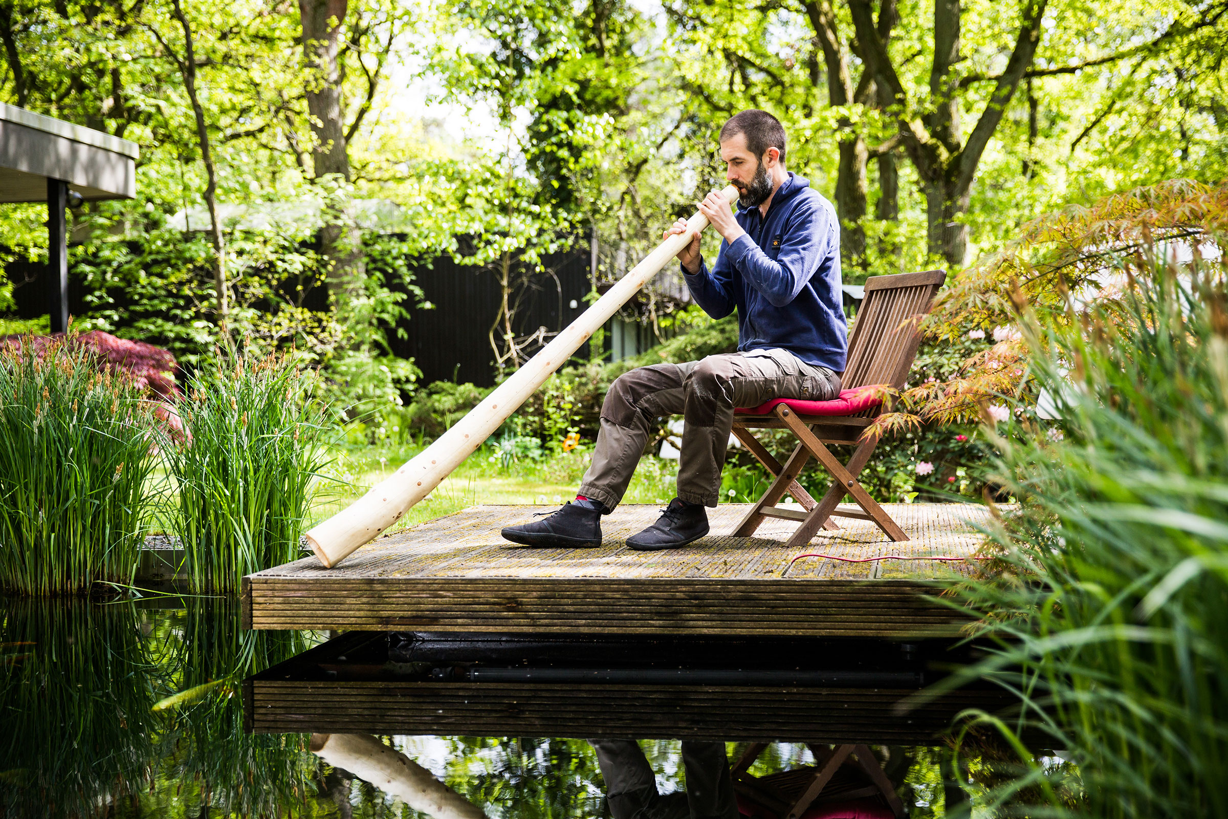 Un homme est assis sur une terrasse en bois et joue du didgeridoo au-dessus d'un étang dans un jardin.