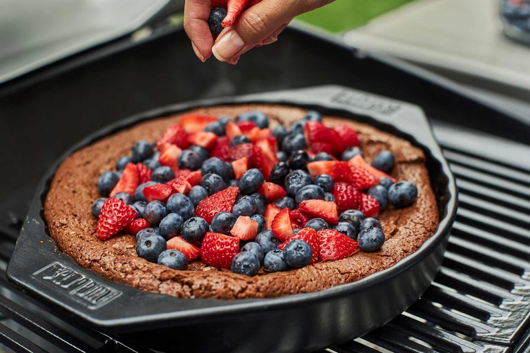 Ein dunkler Kuch an enger Grillform mat Äerdbieren a Blaubeeren gëtt vun enger Hand dekoréiert.