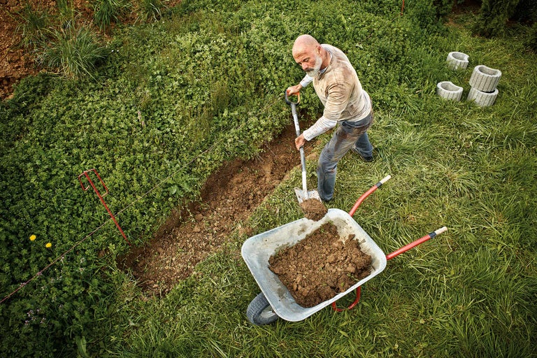 Un homme creuse une tranchée avec une bêche et remplit une brouette dans un jardin.
