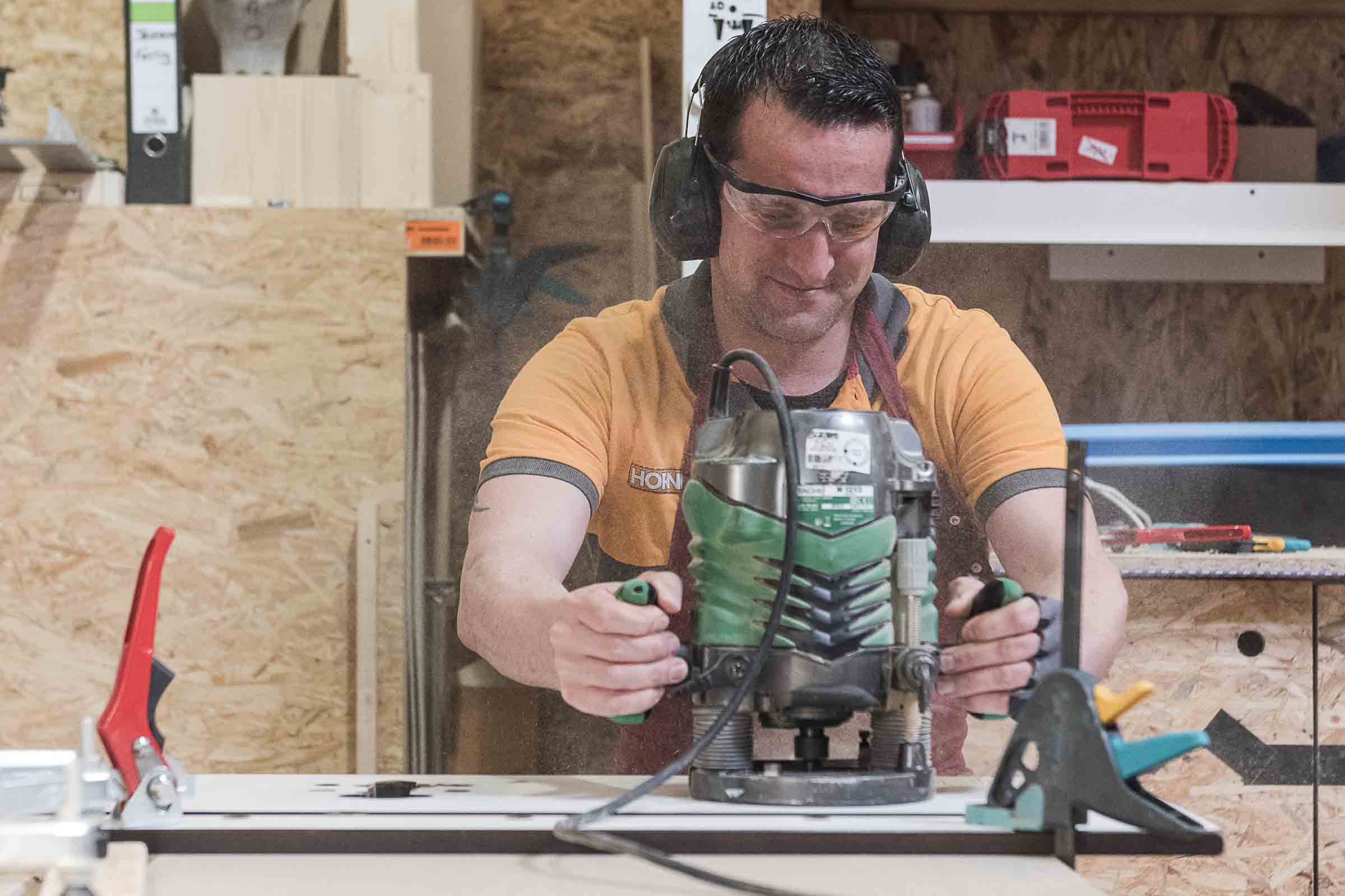 Un homme utilise une défonceuse sur une surface en bois, protégé par des lunettes et un casque antibruit.