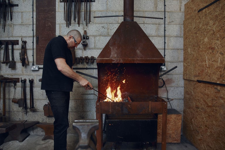 Ein Mann arbeitet mit einem Schmiedeofen in einer Werkstatt. Der Ofen steht im Mittelpunkt des Bildes, während der Mann links davon steht und mit einem Werkzeug im Feuer hantiert. Im Hintergrund sind Werkzeuge an der Wand zu sehen.