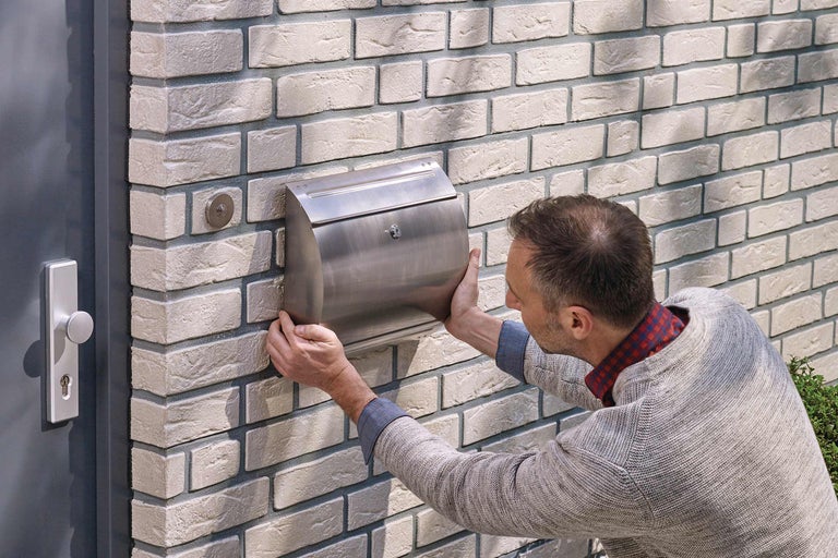 Un homme installe une boîte aux lettres en acier sur un mur de briques à côté d'une porte d'entrée.