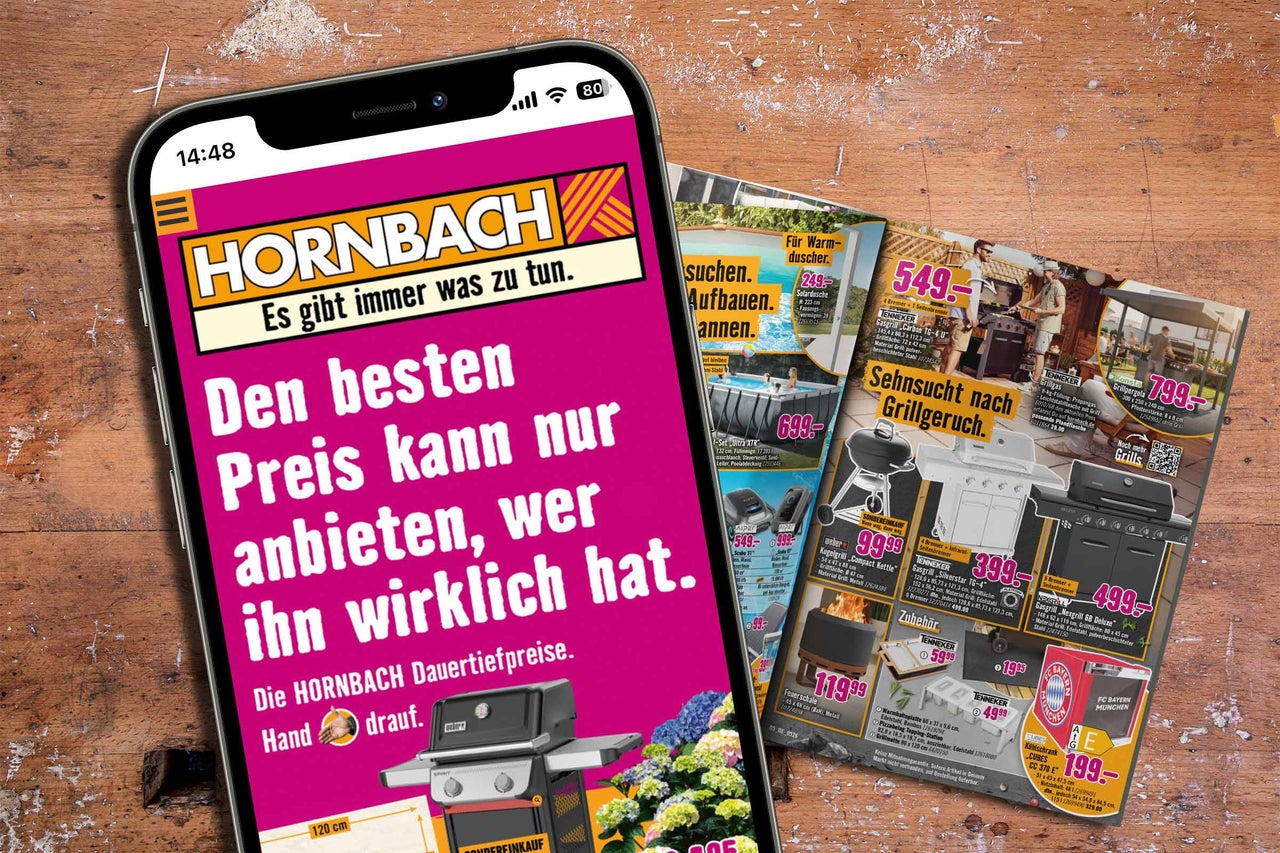 Un smartphone avec l'application Hornbach affichant une publicité pour les prix bas permanents sur une table en bois à côté de catalogues.