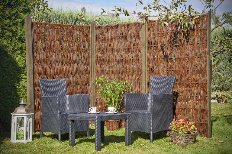 Scène de jardin avec clôture en osier, chaises en rotin et table sur une pelouse.