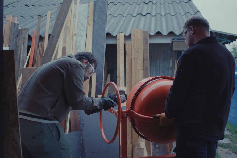 Deux personnes travaillent à l’extérieur avec une bétonnière. L’une utilise une meuleuse d’angle, tandis que l’autre maintient la machine.