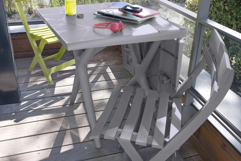 Scène de jardin avec table pliante grise, chaise pliante verte et accessoires sur une terrasse.