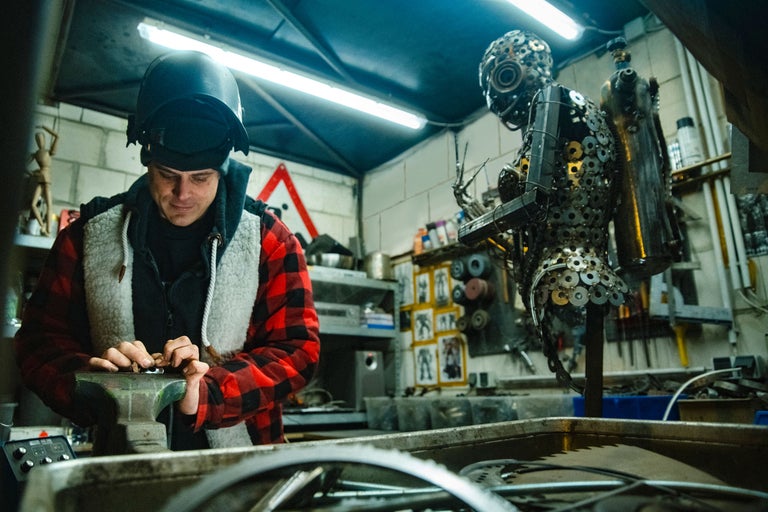 Ein Mann mit Schweißhelm arbeitet in einer Werkstatt an einem Schraubstock, im Hintergrund eine Skulptur.