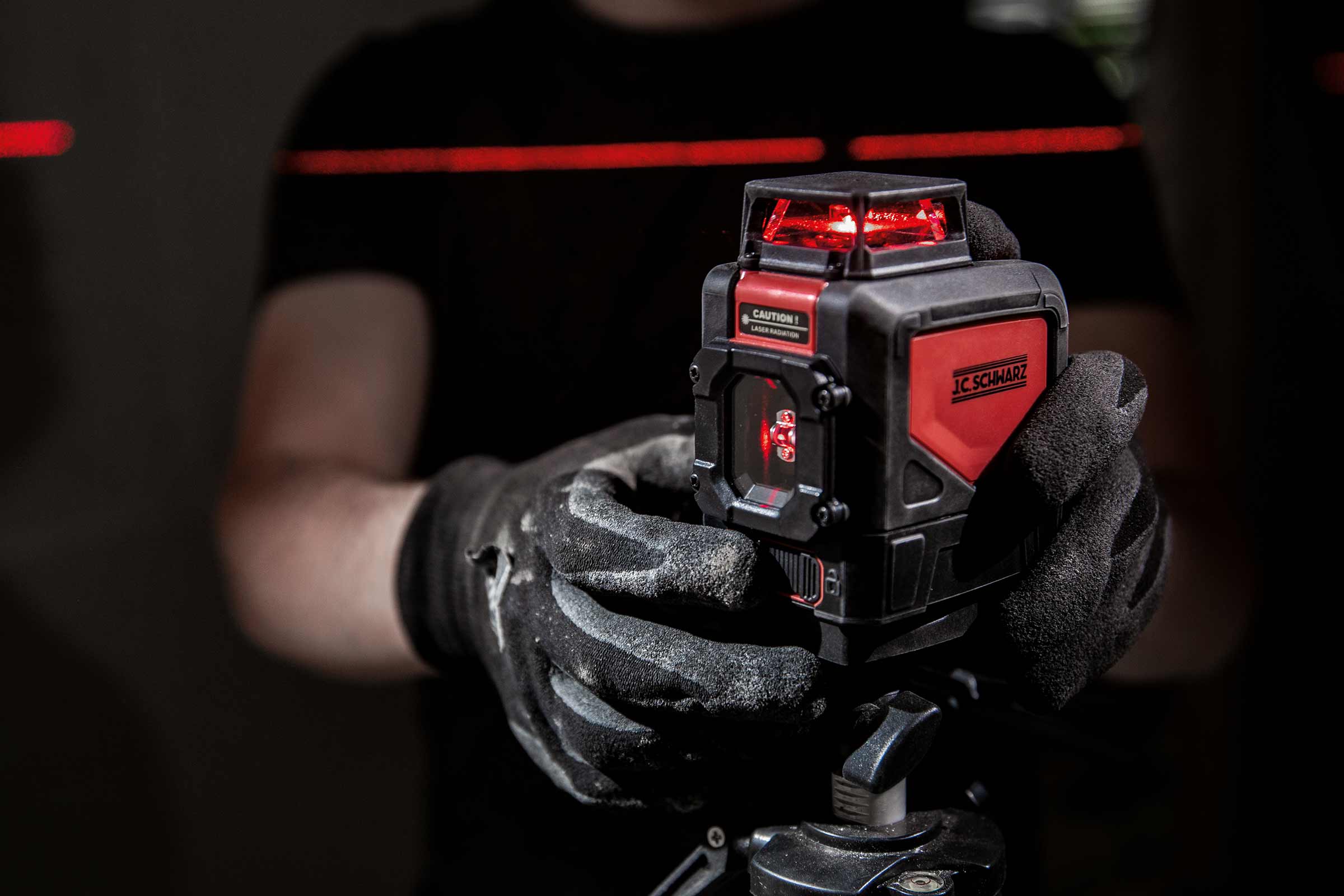 Une personne portant des gants de travail tient un laser lignes croisées J.C. Schwarz sur un trépied, qui projette des lignes laser rouges.