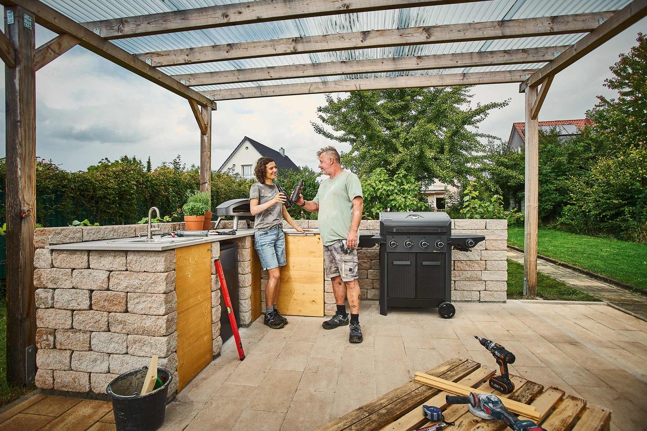 Cuisine d'extérieur en pierre avec barbecue à gaz, four à pizza et évier sous une pergola en bois, deux personnes et outils sur la terrasse.
