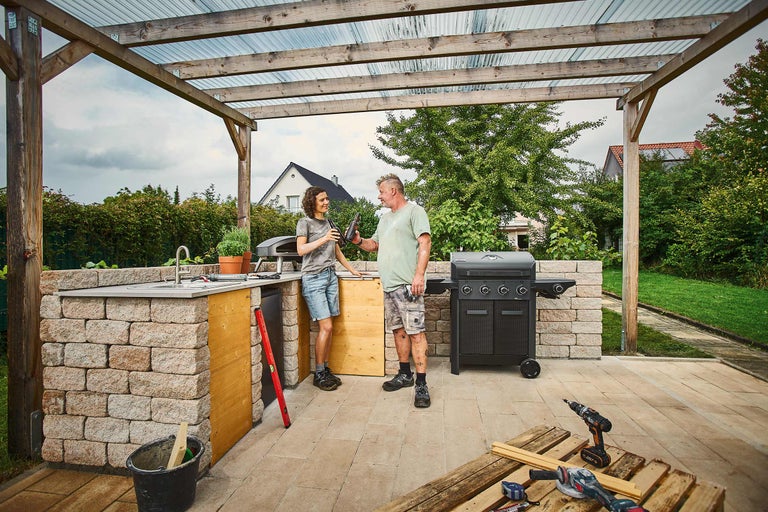 Cuisine d'extérieur en pierre avec barbecue à gaz, four à pizza et évier sous une pergola en bois, deux personnes et outils sur la terrasse.