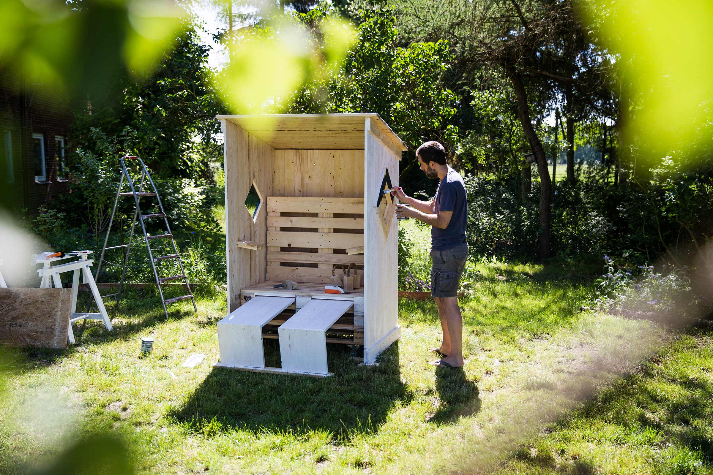 Mann streicht ein Gartenhaus aus Holz im Garten. Im Hintergrund sind Bäume und eine Leiter zu sehen.