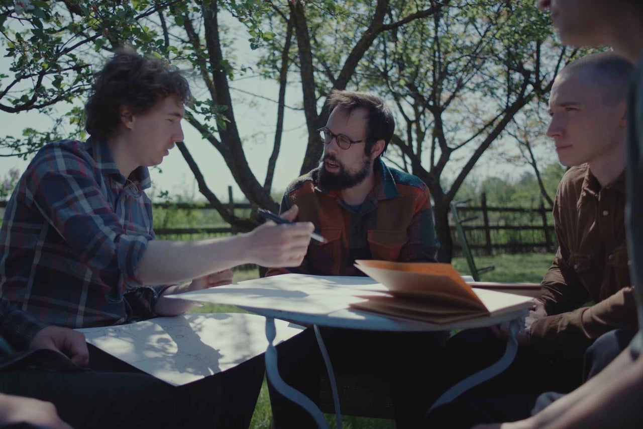 Trois hommes sont assis dehors sous des arbres autour d une table pour discuter de plans de projet.