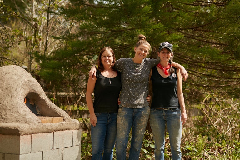 Drei Frauen stehen vor einem Lehmofen im Freien.