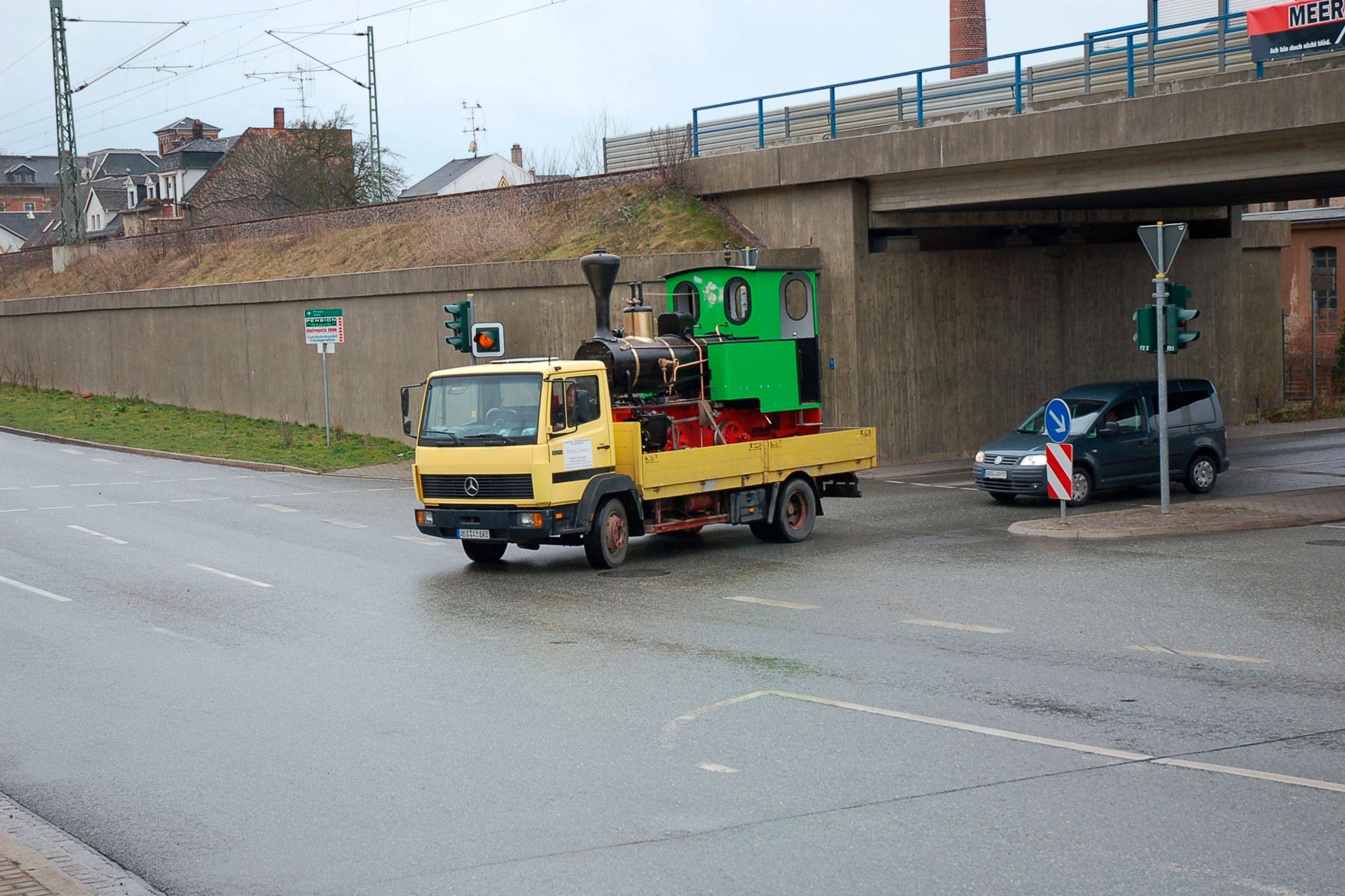 Ein gelber LKW transportiert eine grüne Miniatur Dampflokomotive auf einer öffentlichen Straße.