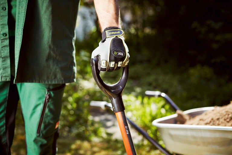 Une personne portant des gants de travail tient une bêche avec une brouette remplie en arrière-plan.