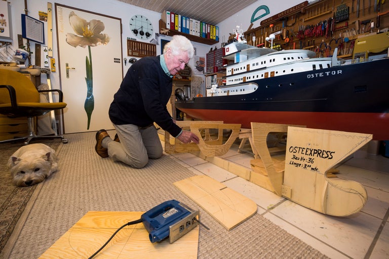 Mann arbeitet an einem Schiffsmodell mit einer Stichsäge in einer Werkstatt, im Hintergrund befindet sich ein Hund