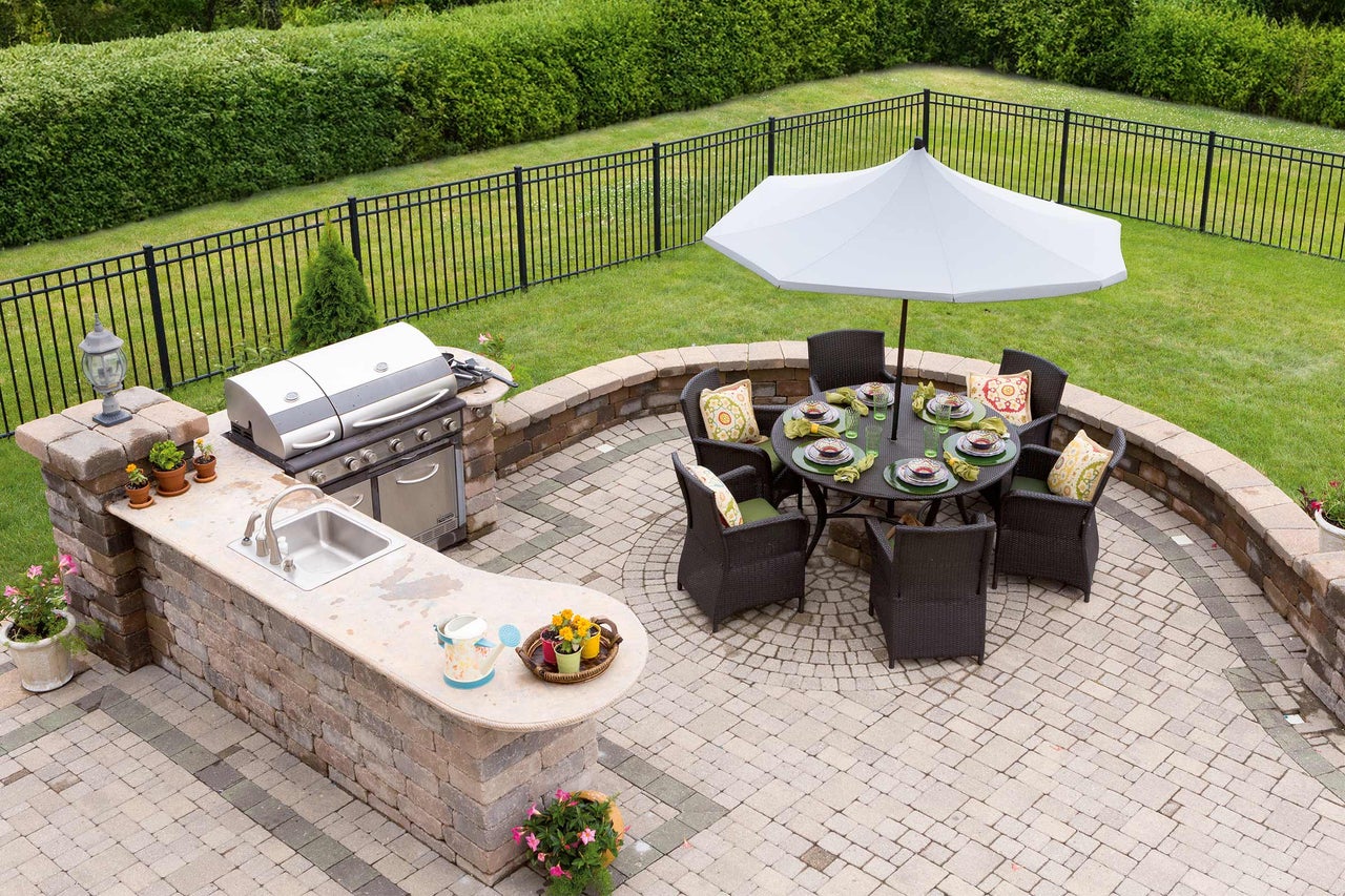 Gartenszene mit Grill, Spüle, Esstisch mit Sonnenschirm und Sitzgelegenheiten auf einer Steinterrasse