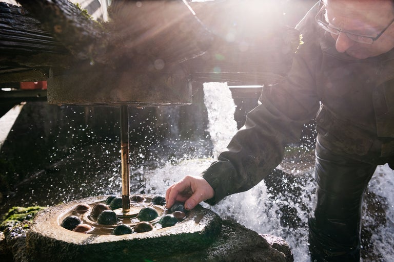 Ein Mann befüllt einen Steinbrunnen mit Flusssteinen unter einem Wasserrad.