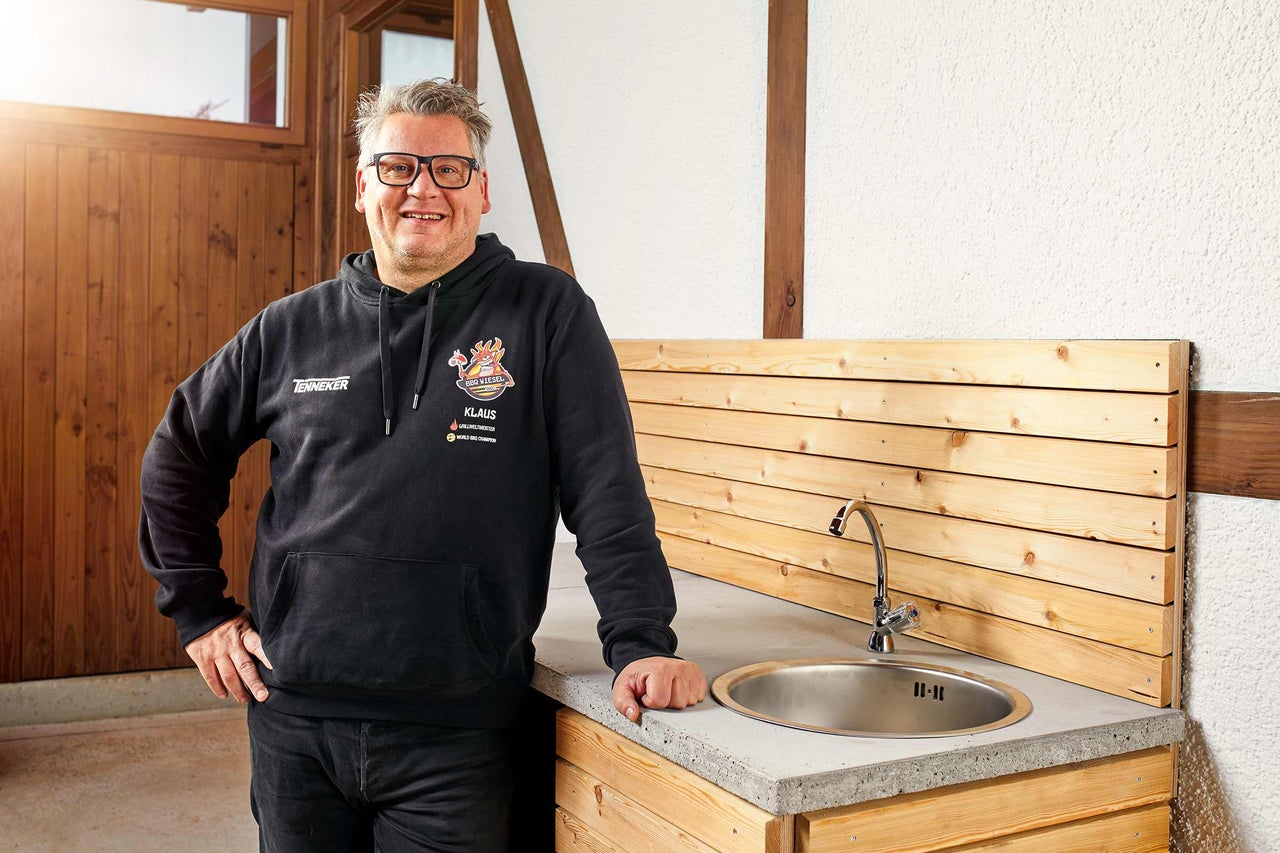 Un homme se tient à côté d'une cuisine extérieure avec évier et mur en bois