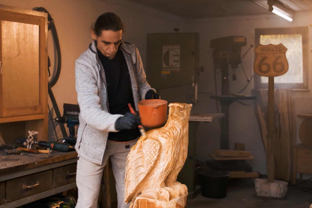 Ein Mann bemalt eine hölzerne Eule in einer Werkstatt. Im Hintergrund Werkzeuge und eine Bohrmaschine.