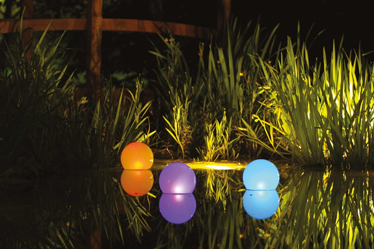 Trois boules flottantes colorées illuminées sur un étang entre des plantes.