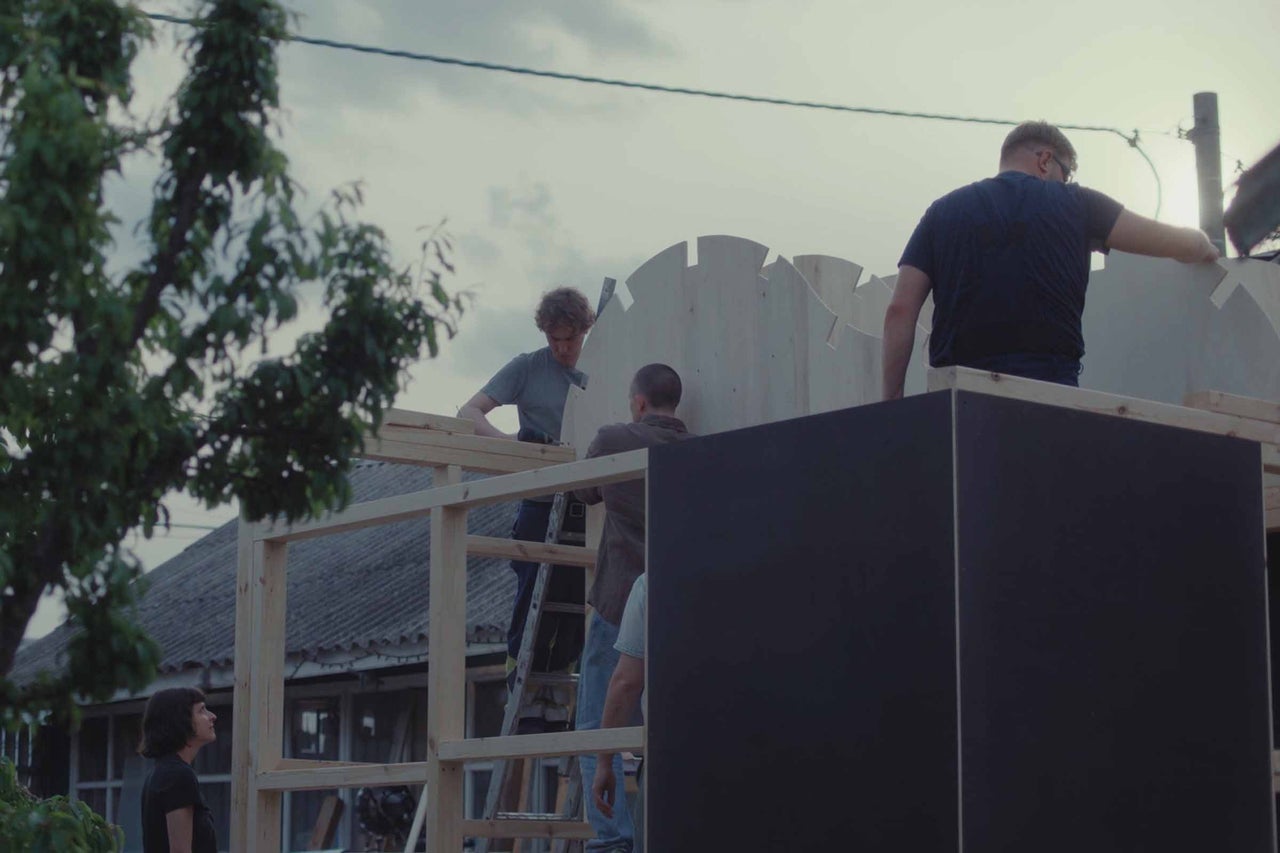 Trois personnes construisent ensemble une maison en bois dans le jardin en fixant les panneaux muraux sur la structure en bois.
