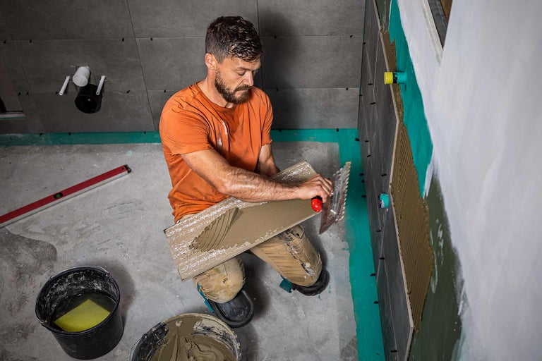 Un carreleur applique de la colle sur un carreau avec une spatule crantée pour le fixer au mur.