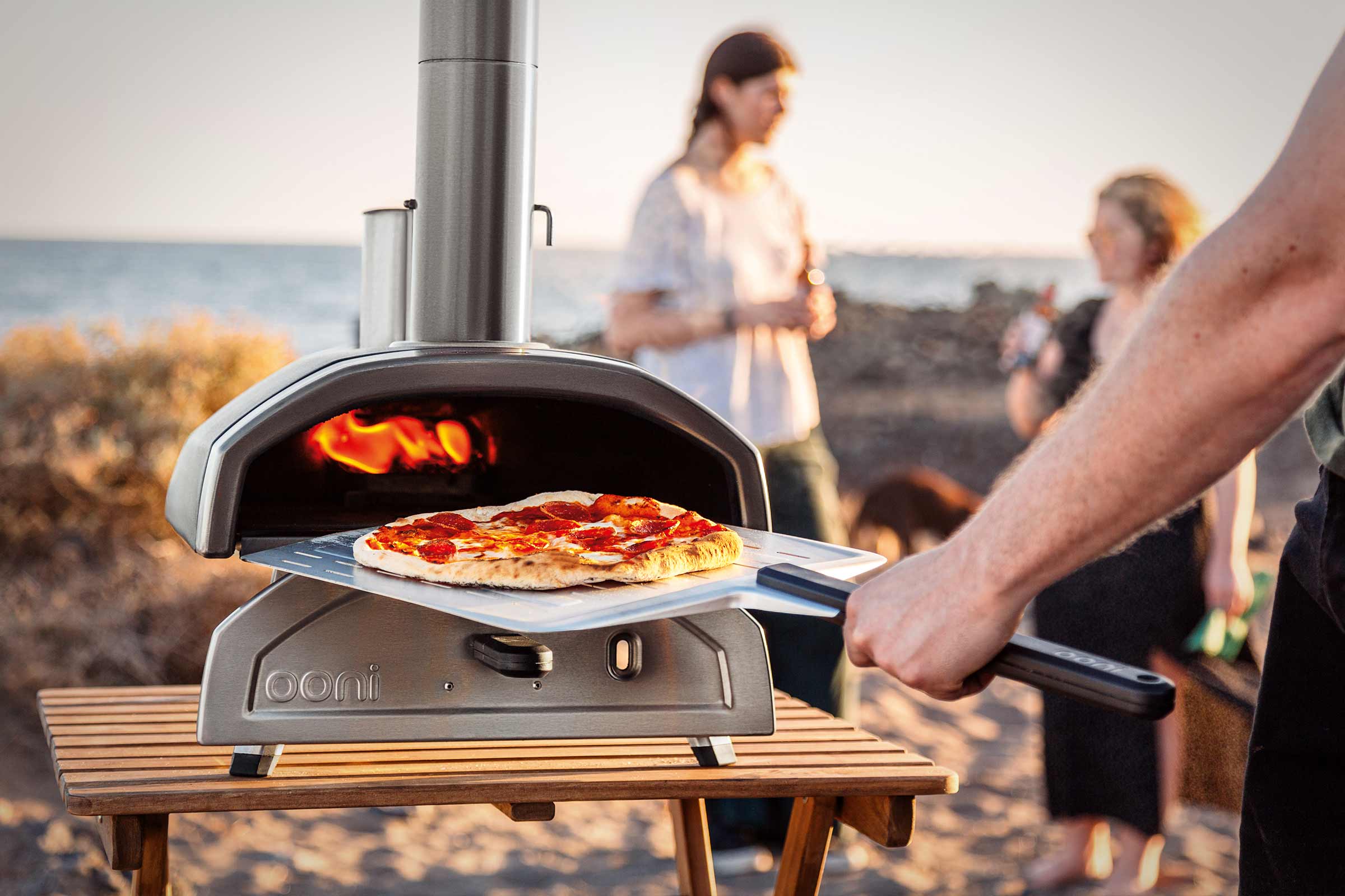 Ooni Pizzaofen mit Pizzaschieber auf einem Holztisch, im Hintergrund Personen am Strand.