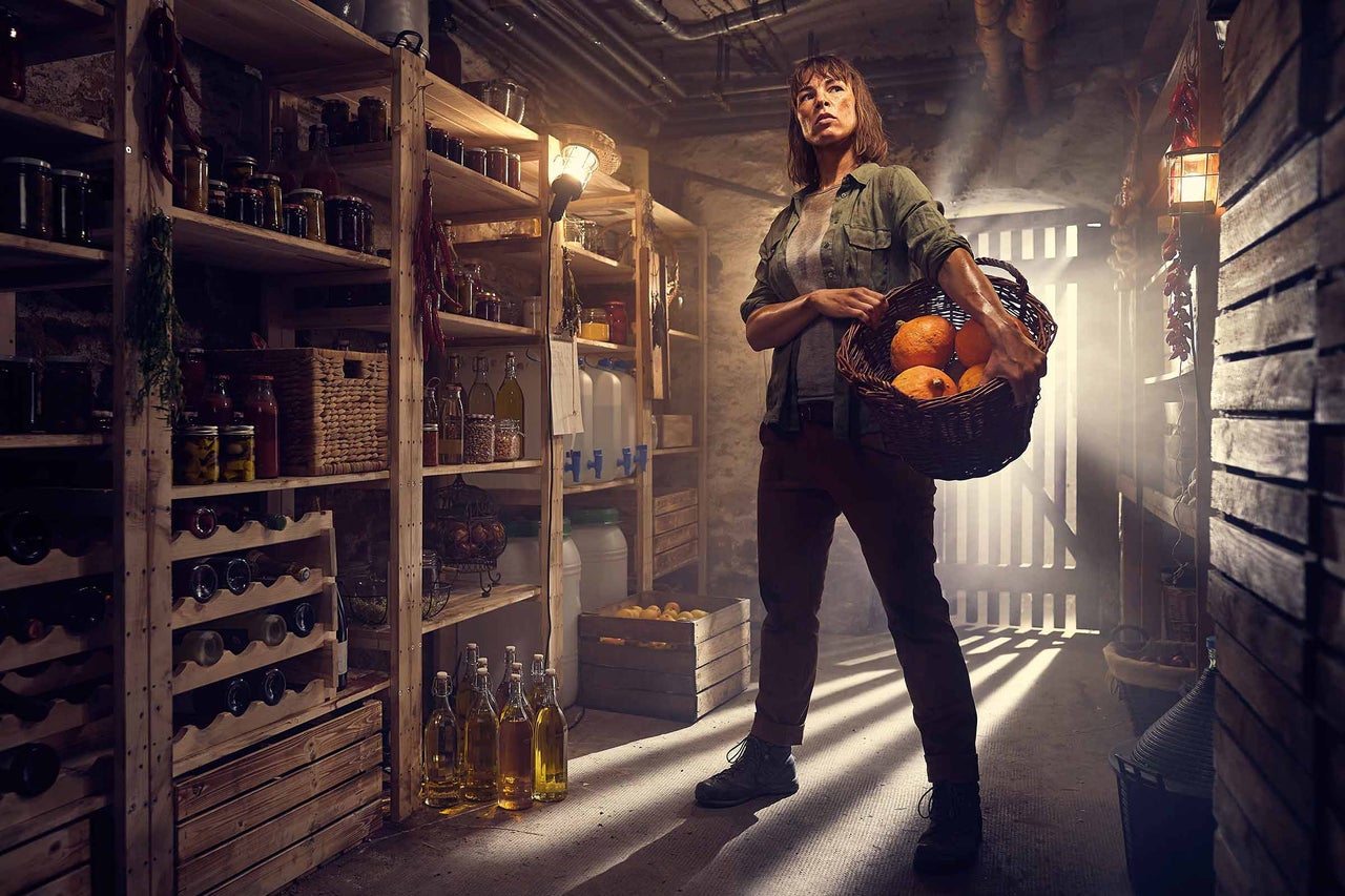 Une femme se tient dans une cave avec des étagères en bois remplies de provisions et tient un panier de citrouilles.