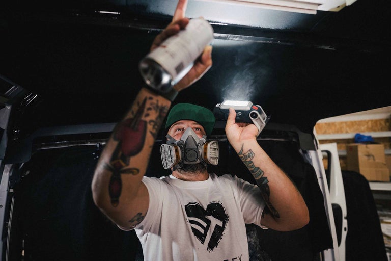 Ein Mann mit Atemschutzmaske sprüht Farbe auf eine Oberfläche, die mit einer Arbeitsleuchte beleuchtet wird