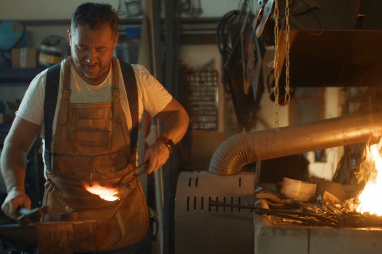 Ein Mann mit Lederschürze schmiedet Metall in einer Werkstatt mit Esse