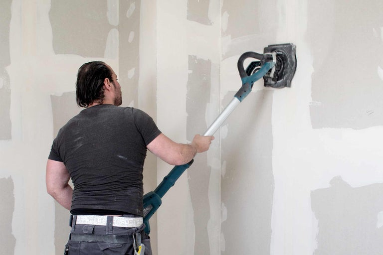 Handwerker schleift eine Wand mit einem Langhalsschleifer. Der Raum wirkt hell und staubig.