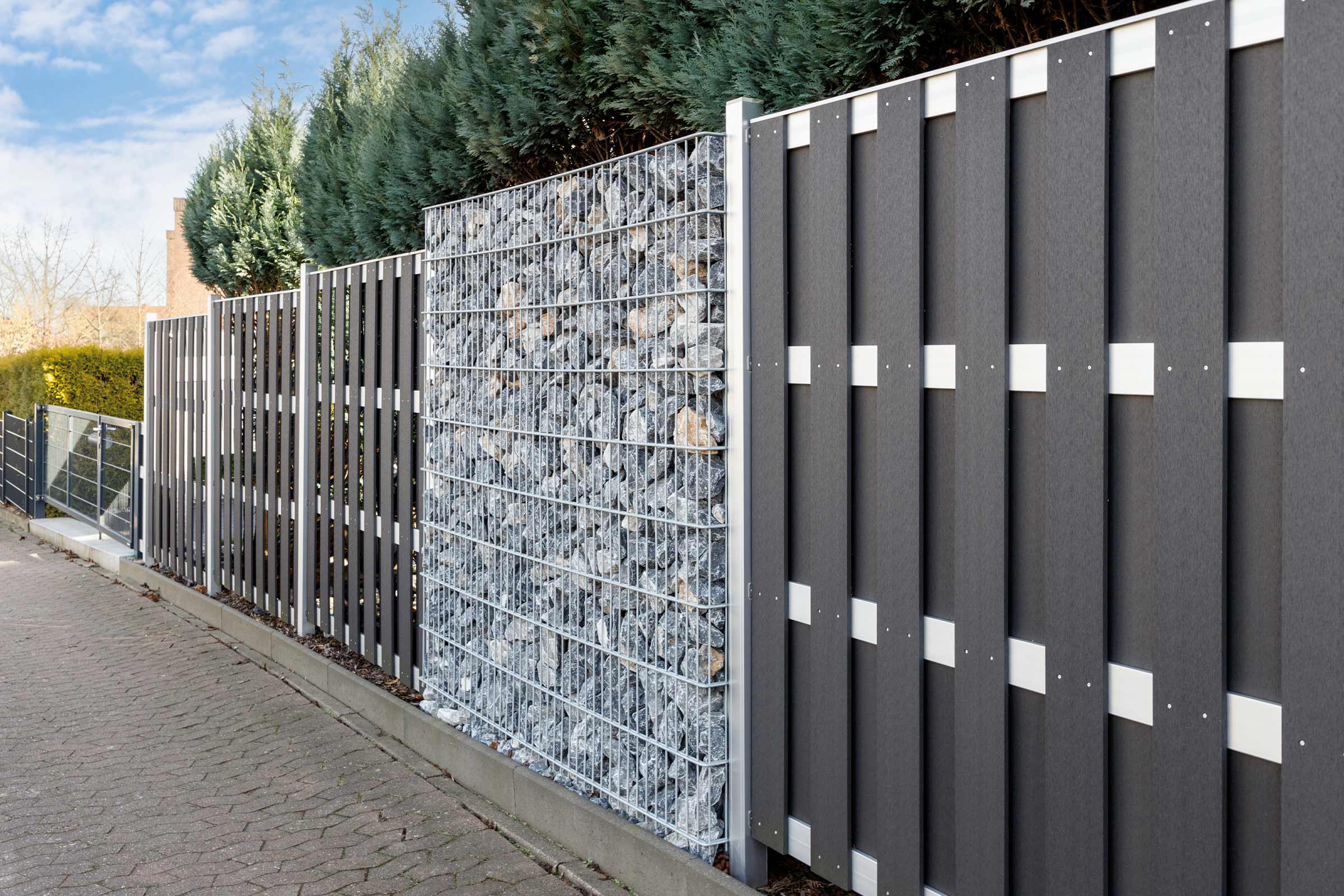 Verschiedene Zaunelemente aus Kunststoff und Gabionen bilden eine Grundstücksbegrenzung.