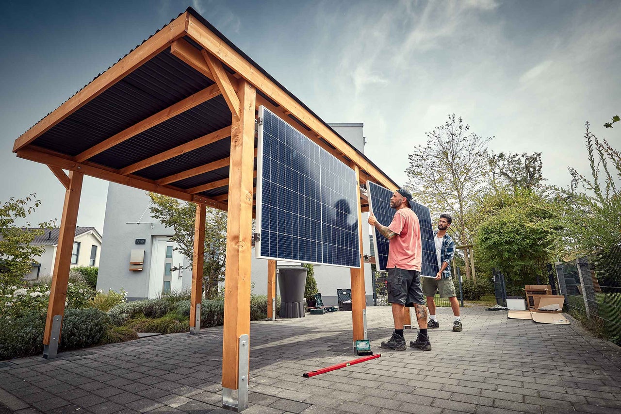 Deux personnes installent des panneaux solaires sur un abri de voiture en bois avec un toit en tôle ondulée devant une maison.