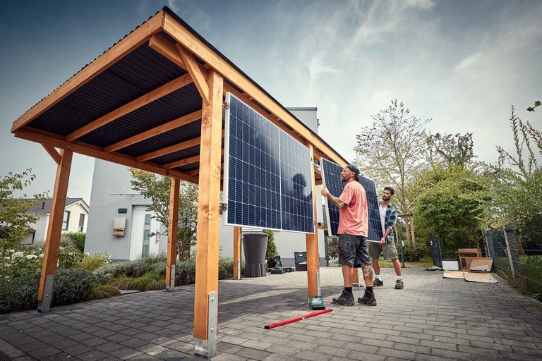 Deux personnes installent des panneaux solaires sur un abri de voiture en bois avec un toit en tôle ondulée devant une maison.
