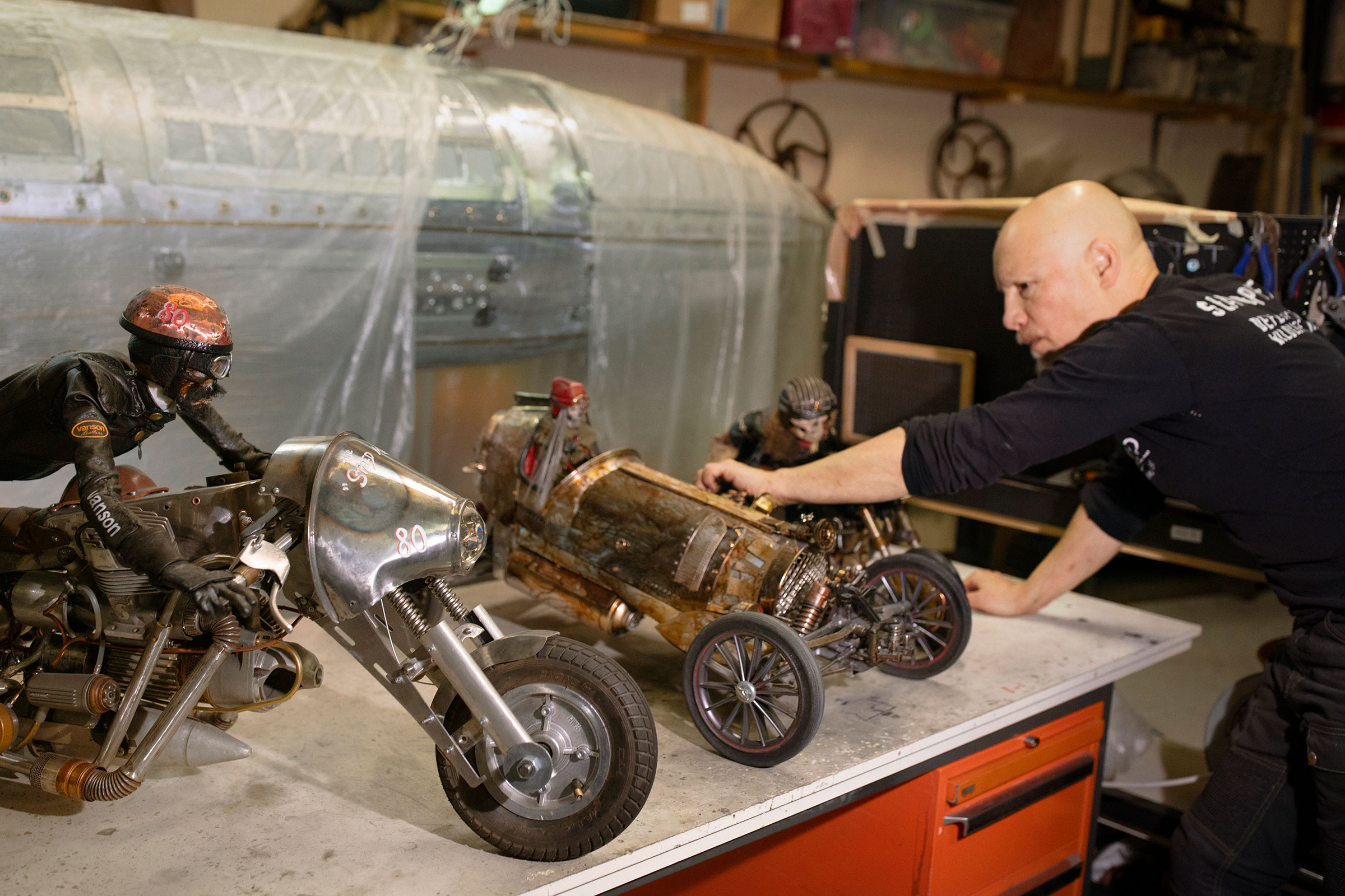 Mann präsentiert Modellmotorrad und Modellrennwagen aus Metall auf einer Werkbank.