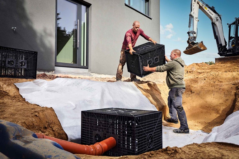 Zwei Männer installieren Rigolenblöcke für die Regenwasserversickerung in einer Baugrube, ausgelegt mit einem weißen Vlies.