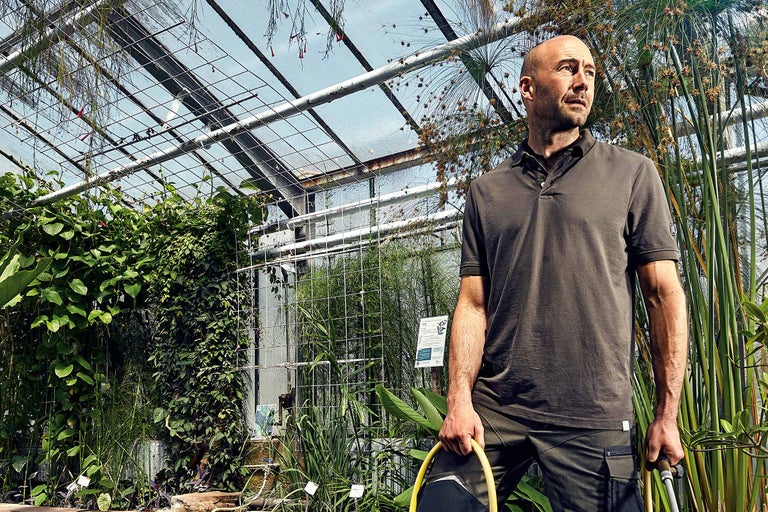 Homme portant un polo et un pantalon dans une serre avec des plantes et un tuyau d'arrosage