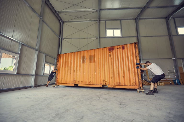 Deux personnes déplacent un conteneur de stockage sur roulettes dans un entrepôt à structure métallique.
