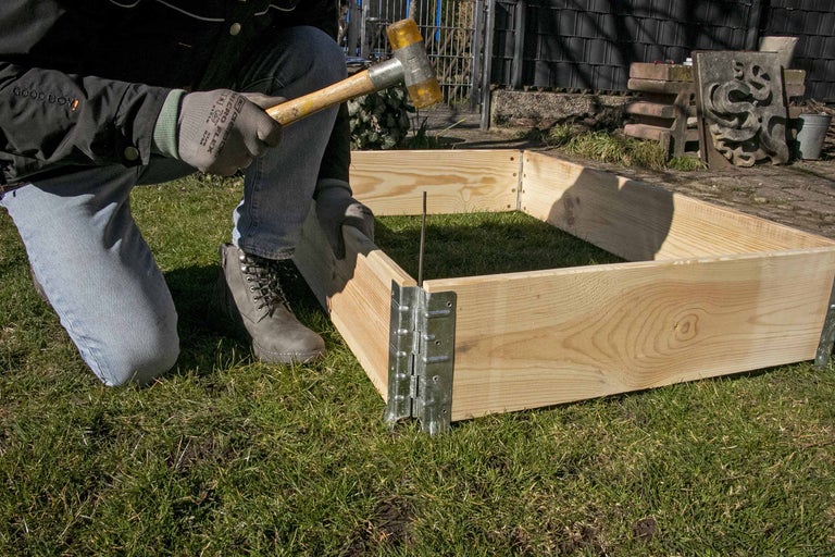 Une personne assemble un potager surélevé en bois avec un marteau et des équerres dans le jardin.