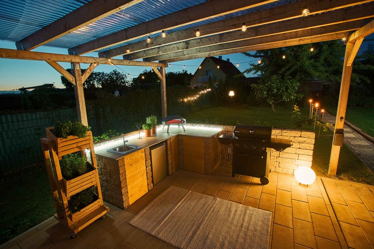 Beleuchtete Outdoor-Küche mit Grill, Spüle und Pflanzregal unter einer Pergola mit Lichterkette