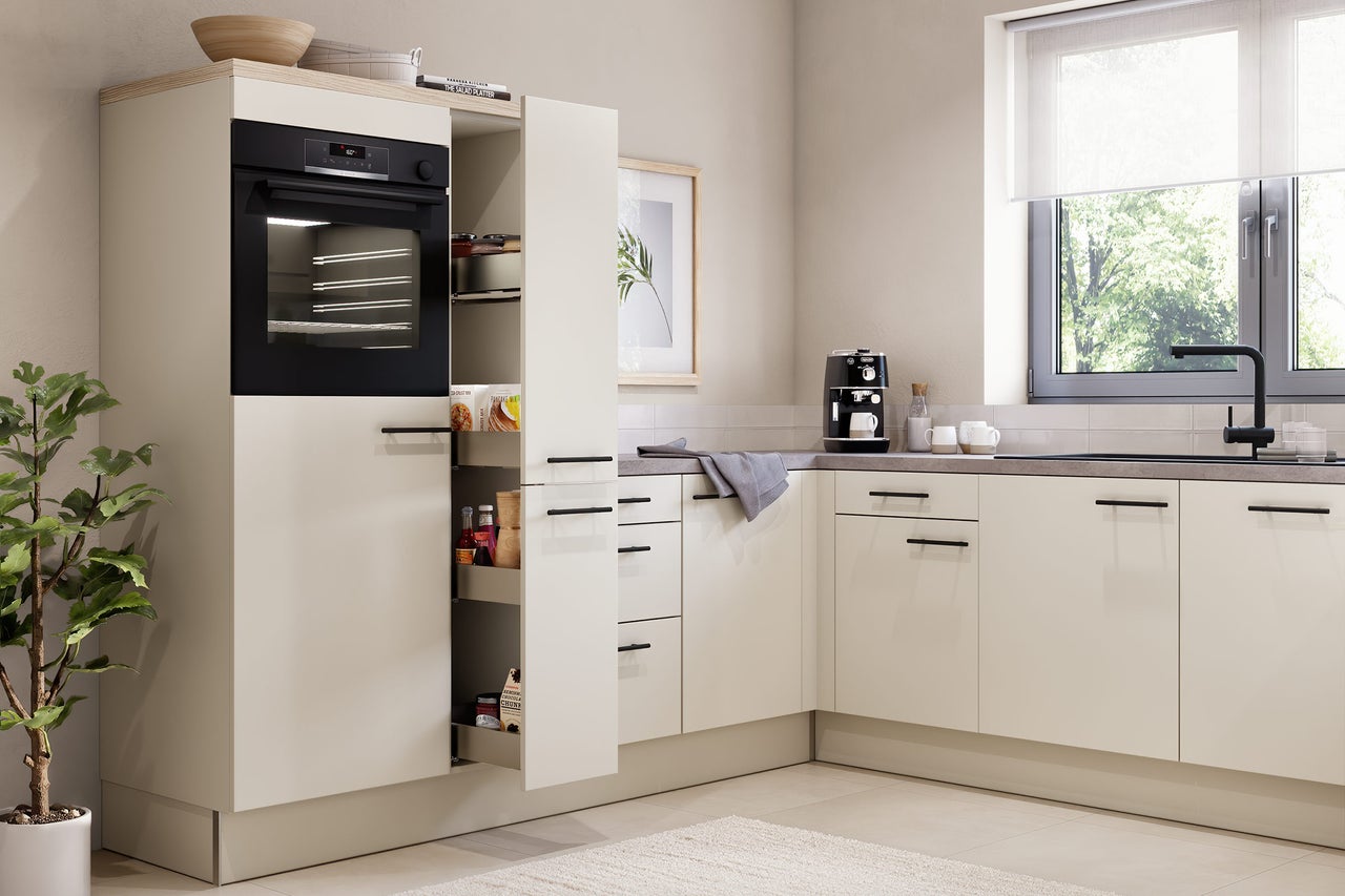 Cuisine moderne en L de couleur beige avec four encastré noir, armoire à pharmacie, plan de travail gris et robinet noir près de la fenêtre.