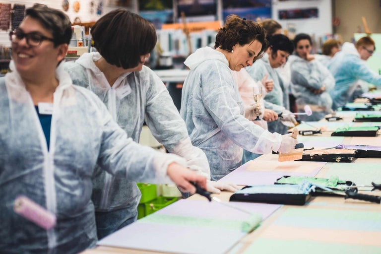 Plusieurs personnes travaillent avec de la peinture, des pinceaux et des rouleaux sur des tables dans une pièce lumineuse.