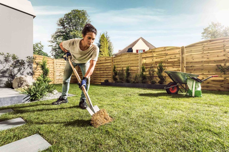 Femme travaillant avec une bêche dans le jardin, une clôture en bois et une brouette en arrière-plan